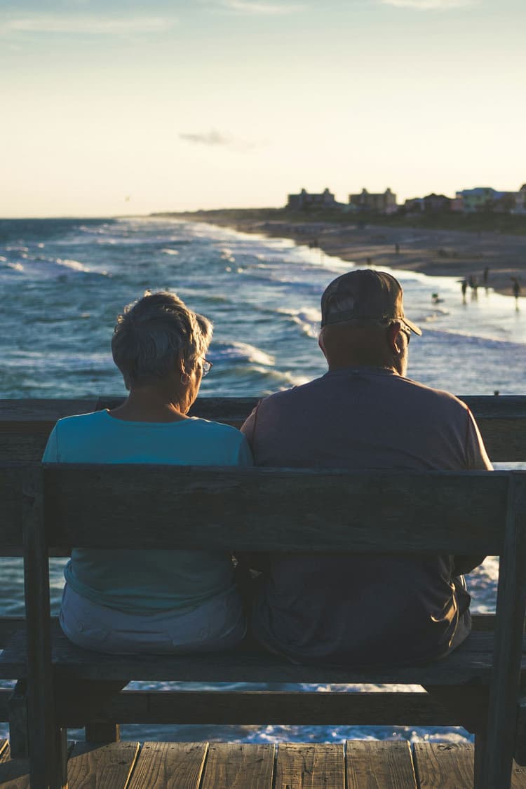 Retired couple sitting on bench looking at ocean.